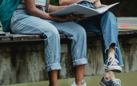 Crop faceless diverse schoolgirls reading textbook on pier in park